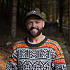 Smilende mann i mønstret strikkegenser og caps foran trær i en skog.