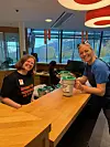 To smilende kvinner ved et trebord i en lys kafé med drikkevarer.