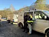 To personer ved en hvit varebil og tilhenger på en grusplass i skoglandskap.