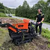 Person ved en oransje beltemaskin på en grusvei i et grønt skogsområde.