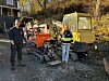 To personer ved anleggsmaskiner, blant annet en gul beltegraver, på et gruslagt område.
