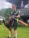 Skogsarbeider med hjelm, sele, tau og motorsag i en hage foran et hus.