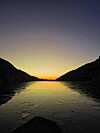 Solnedgang mellom mørke fjell over en stille, delvis islagt fjord.