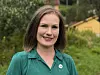 Smilende kvinne i turkis skjorte foran grønt løvverk og et gult hus.