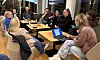 Gruppe voksne i samtale rundt kafébord med kaffe, notater og en bærbar datamaskin.