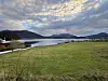 Grønn eng ved en stille fjord, med lave fjell og dramatiske skyer.