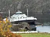 Hvit ferge på fjord ved en liten brygge, omgitt av høstskog og fjell. En lense ligger på vannet rundt ferja.