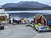 Fergehavn med biler, rødt sjøhus, fjord og fjell i bakgrunnen.
