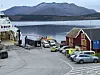 Fergehavn med biler, rødt sjøhus, fjord og fjell i bakgrunnen.