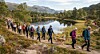 Turgåere går på en sti langs et fjellvann med skog og fjell i bakgrunnen.