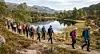Turgåere går på en sti langs et fjellvann med skog og fjell i bakgrunnen.
