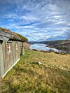 Værbitt trehus i en gresskledd skråning med utsikt over fjord og skogkledde åser.