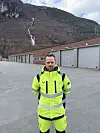 Arbeider i gule verneklær foran garasjebygg med fjell og foss i bakgrunnen.