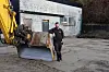 Anleggsarbeider ved skuffen på en gul gravemaskin foran en industribygning.