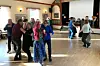 Flere voksne danser i et lyst selskapslokale med tregulv.