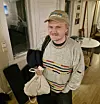 Smilende mann med caps og stripet genser står innendørs med handlepose og ryggsekk.