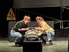 To personer sitter på huk ved en bilstol på en teaterscene.