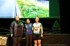 Tre damer står og synger på en scene med treningsklær, ei av de med startnummer.