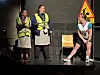 Tre personer på en scene. To av de med refleksvester. Stol og varselskilt i bakgrunnen.
