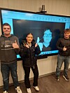 Tre personer med tommel opp foran en stor skjerm som viser et ansikt.