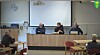 Møte med tre personer ved et panelbord og deltakere på stoler i et møterom.